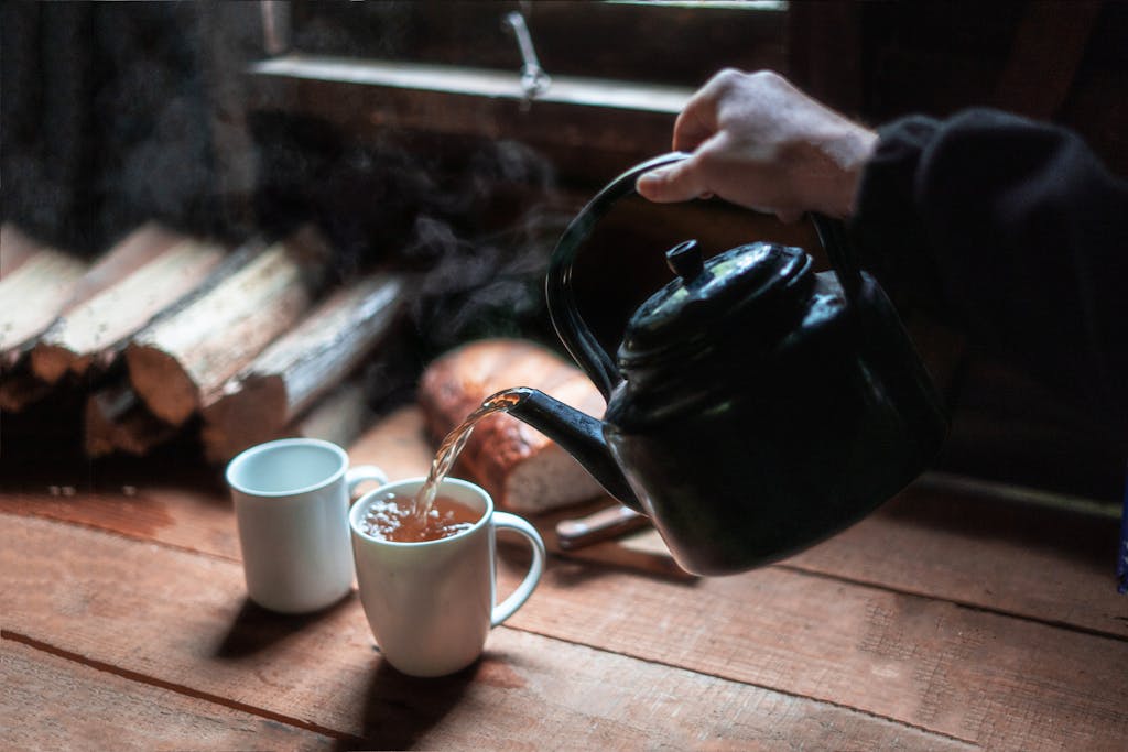 Rustic scene of hot tea being poured from a teapot into a mug by the window.