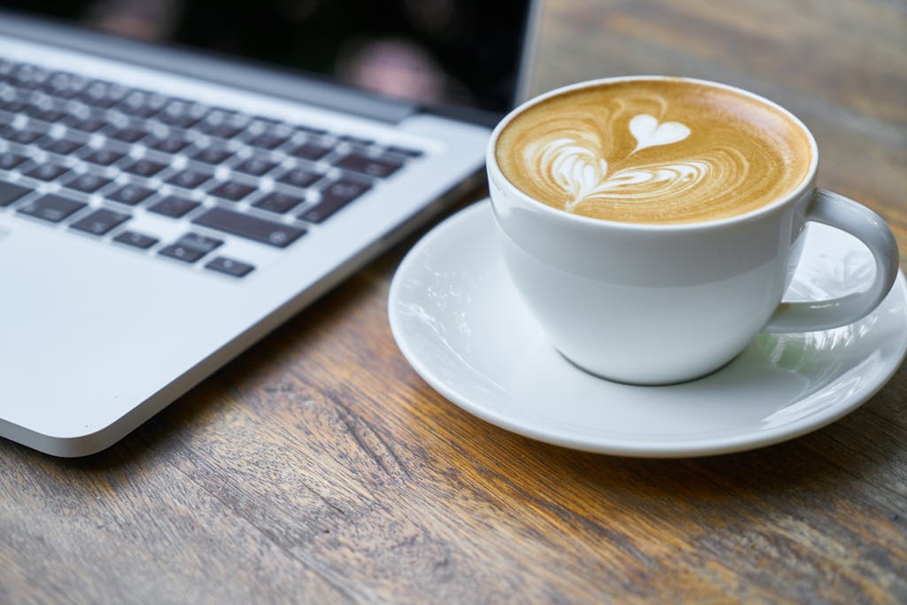 A close-up view of a silver laptop and a cup of latte with heart-shaped latte art on a wooden table. The laptop’s keyboard is visible, and the coffee sits on a white saucer.