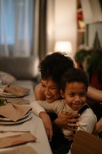 A mother lovingly embraces her son at a warmly lit dining table, capturing a moment of happiness.