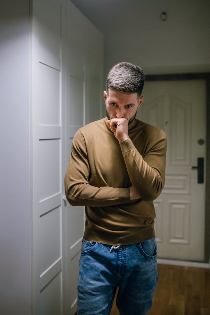 A man in a brown sweater, deep in thought, standing in a hallway with arms crossed, looking pensive.
