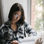 Woman reading a book by the window in a plaid shirt, conveying a peaceful moment of solitude.