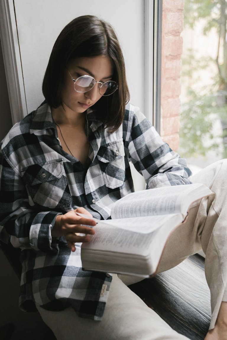 Woman reading a book by the window in a plaid shirt, conveying a peaceful moment of solitude.