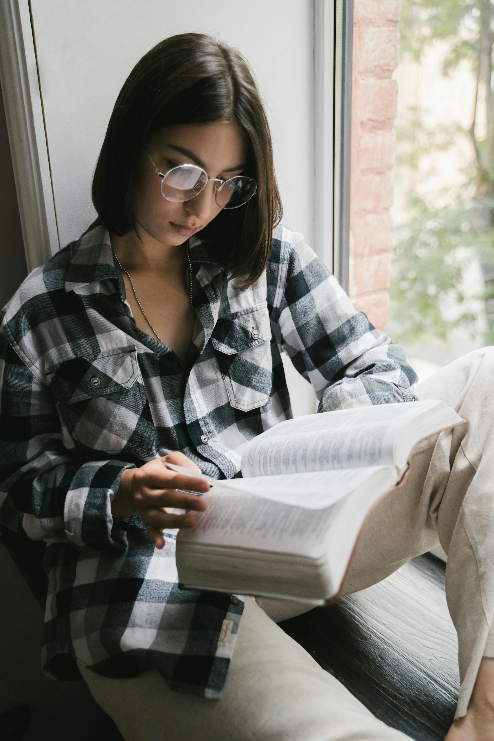 Woman reading a book by the window in a plaid shirt, conveying a peaceful moment of solitude.