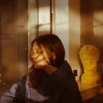 Young woman in a hoodie sitting indoors with artistic shadows, calm and thoughtful.