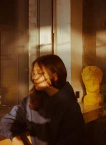 Young woman in a hoodie sitting indoors with artistic shadows, calm and thoughtful.