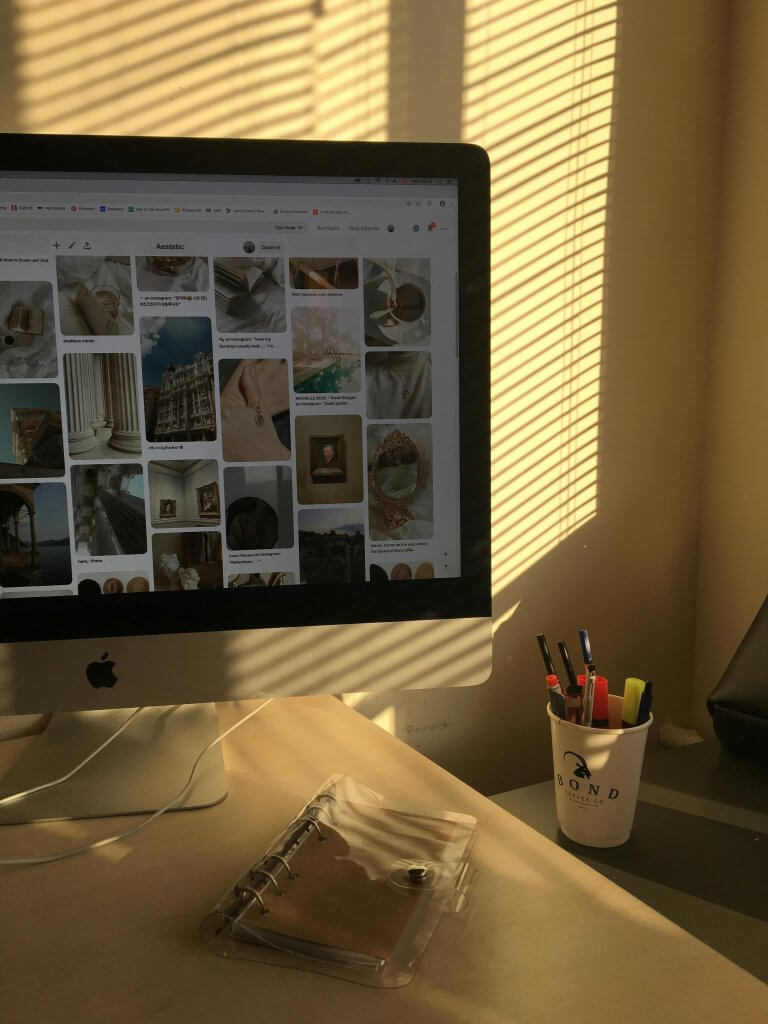 Desk workspace with computer, notebook, and warm sunlight streaming through window blinds.